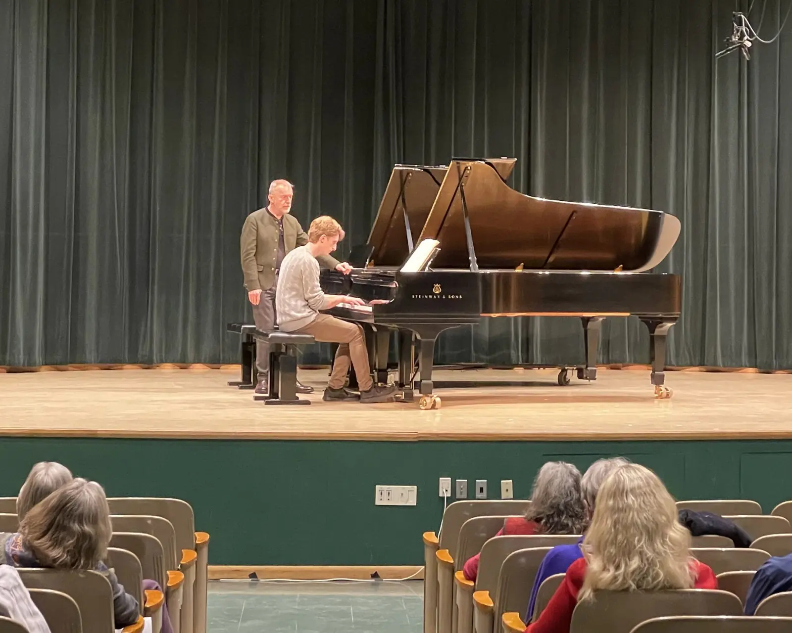 An instructor observes a student playing a grand piano on a stage in front of an audience.
