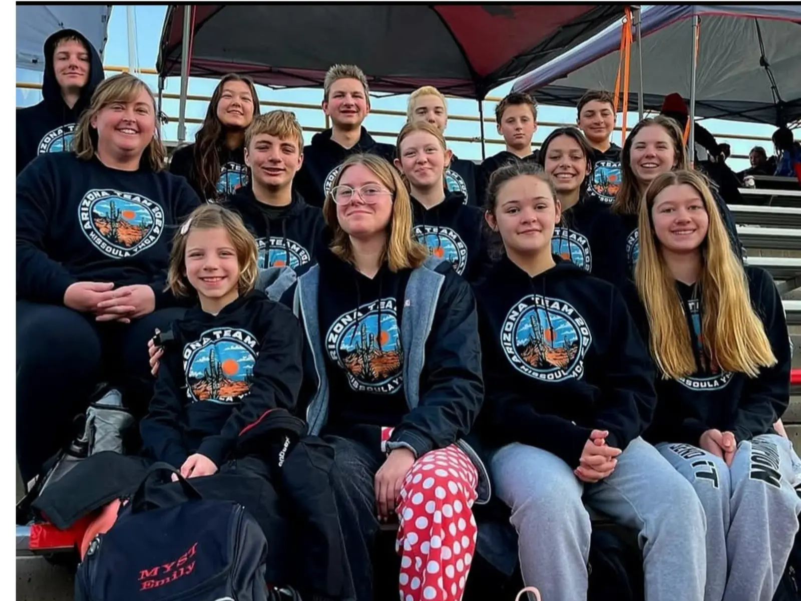 A group of young people and an instructor wearing matching team hoodies sit together in bleachers.