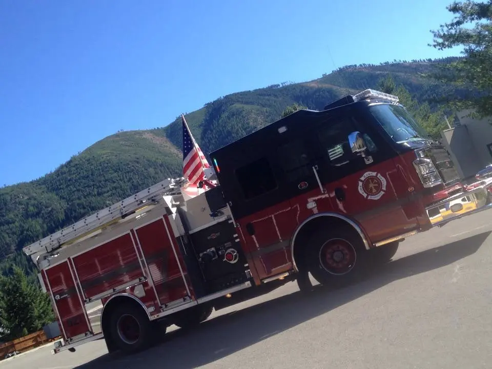 A red and black fire truck with an American flag attached is parked against a backdrop of mountains.
