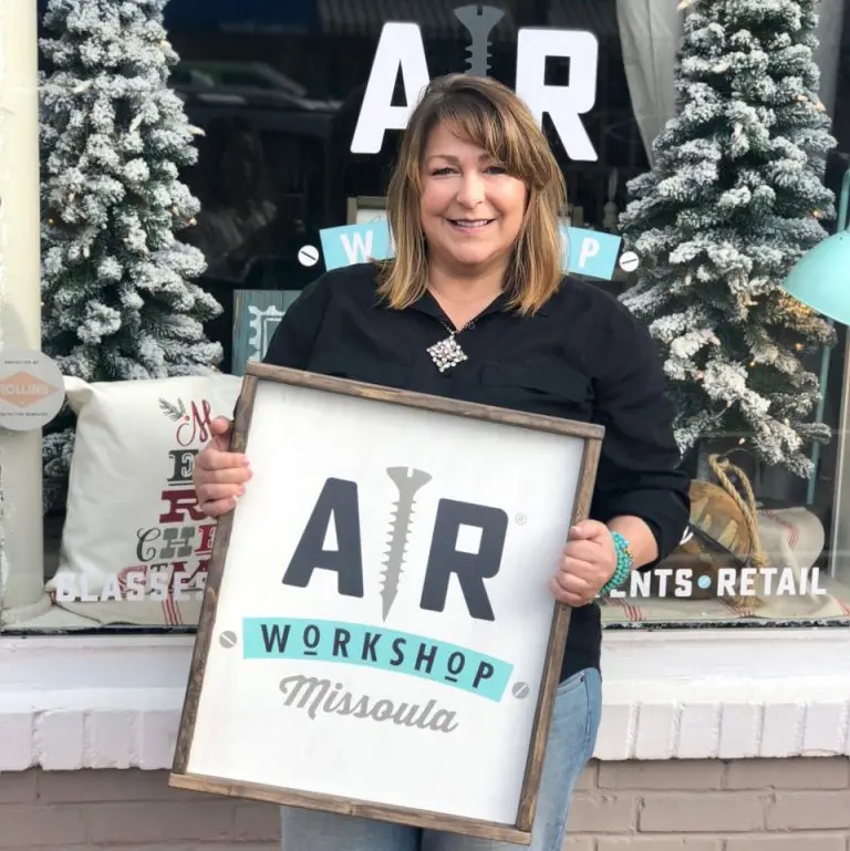 A woman holds a wooden sign with the logo of a business in front of its storefront window.