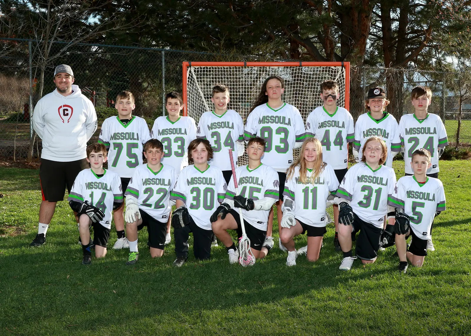 A youth lacrosse team and their coach pose for a group photo in front of a lacrosse goal.