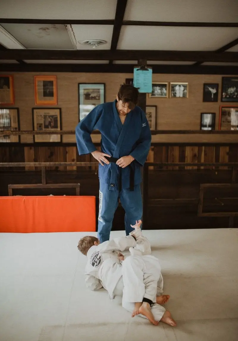 An instructor in a blue gi observes a child practicing on a mat.