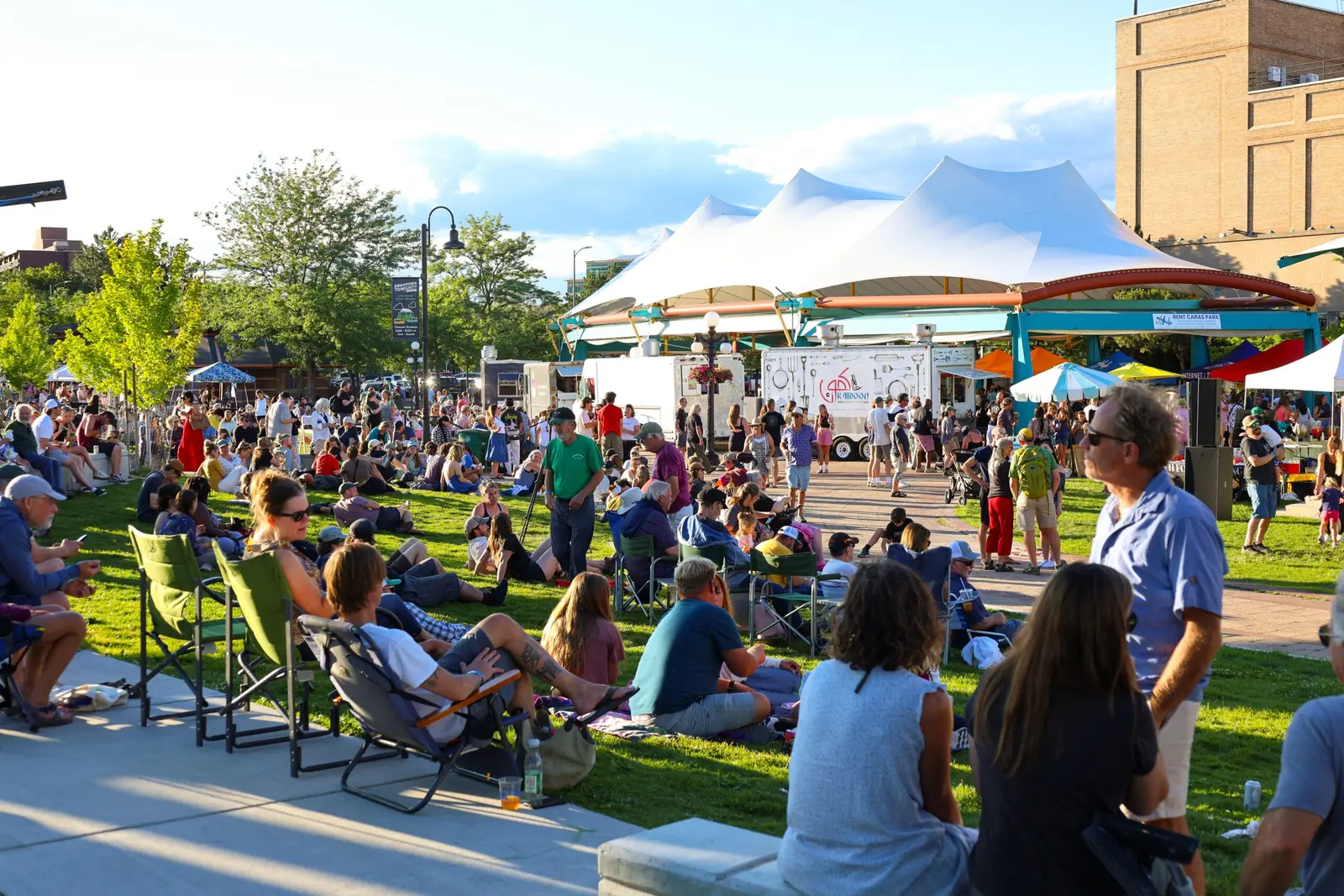 A crowd of people relaxes on a grassy field in front of a large white canopy and food trucks.