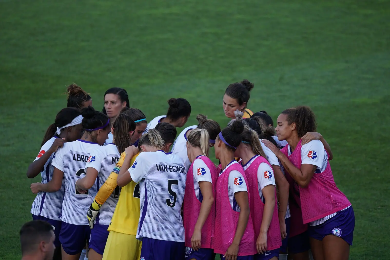 A group of female soccer players stands in a huddle on a grass field.