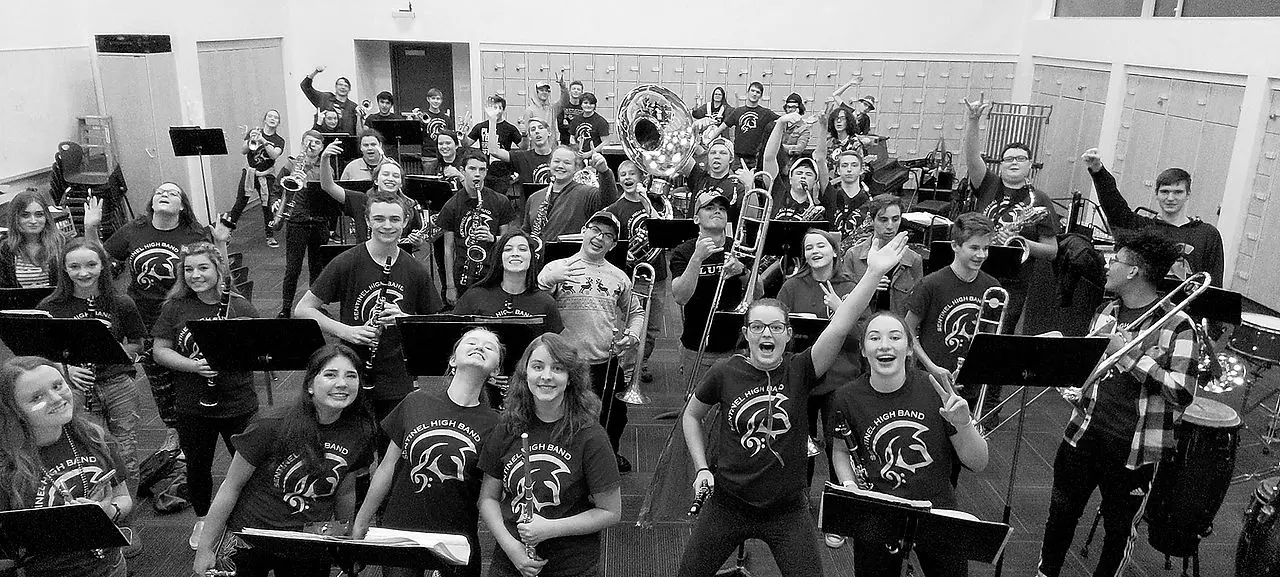 A group of high school band students in matching t-shirts are posing together in a practice room.