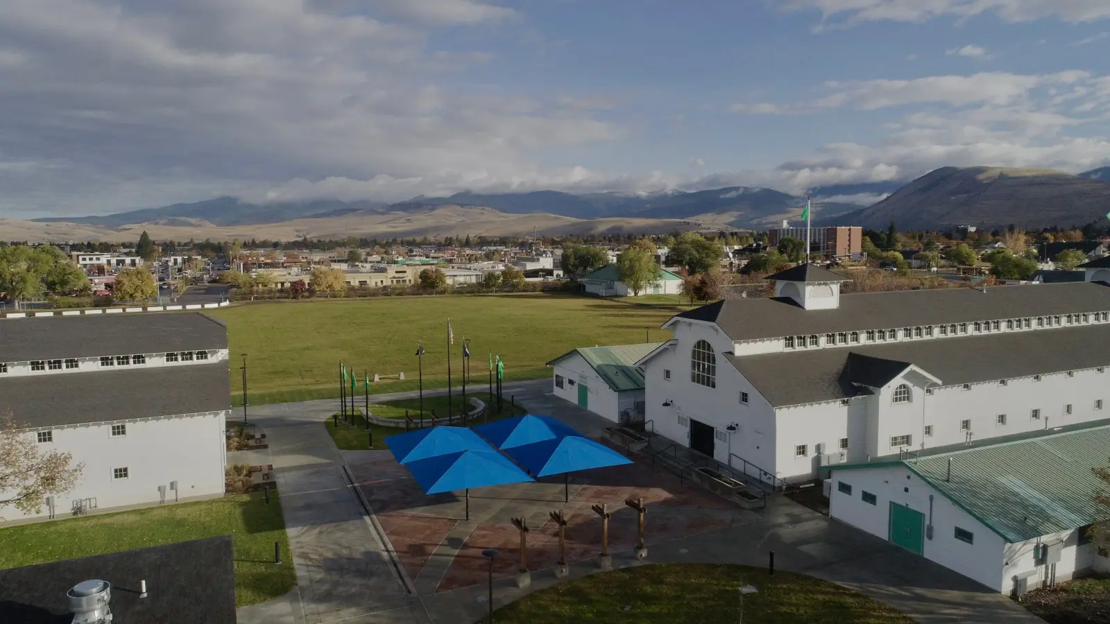 An aerial view shows a complex of white buildings and a central plaza with blue sunshades against a mountain backdrop.