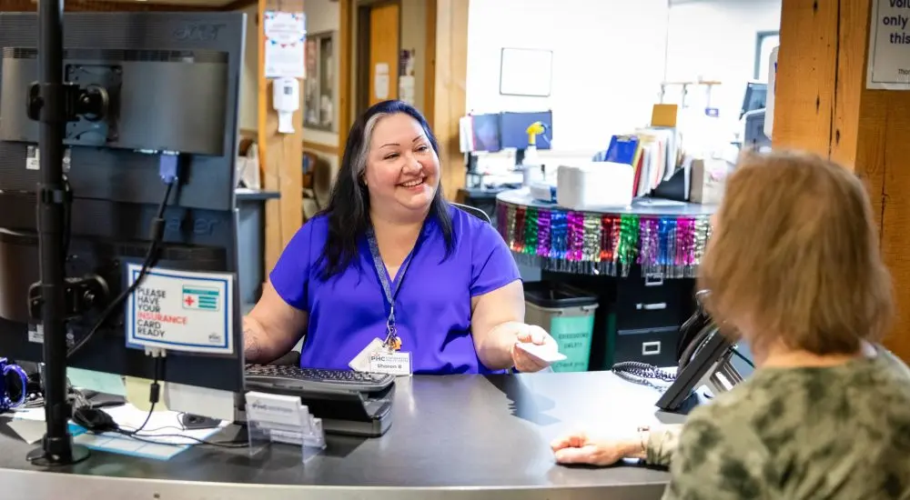 A smiling receptionist at a front desk hands a card to a person standing in front of the counter.