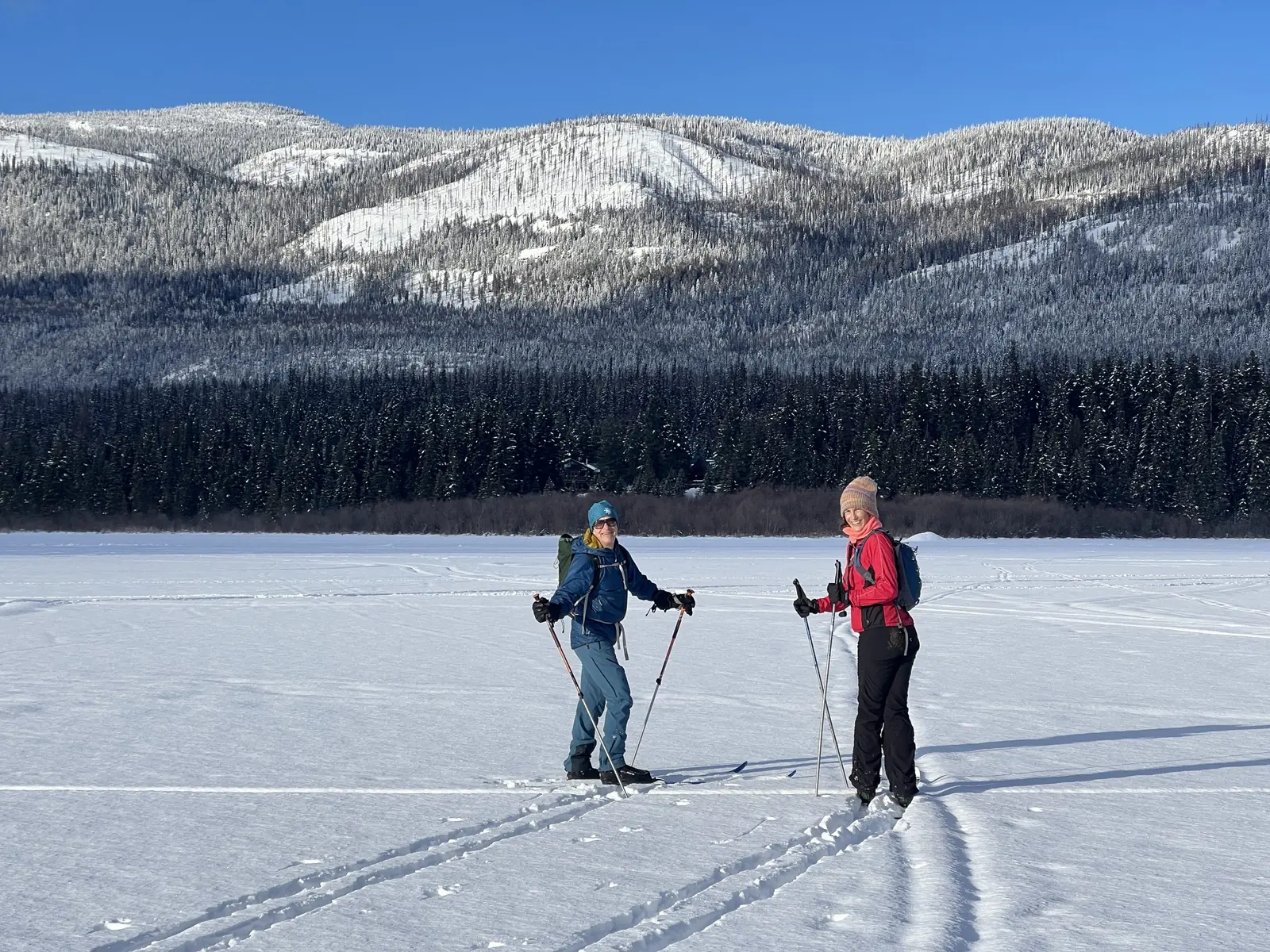 Two people cross-country ski across a wide, snow-covered landscape against a backdrop of a forested mountain range.