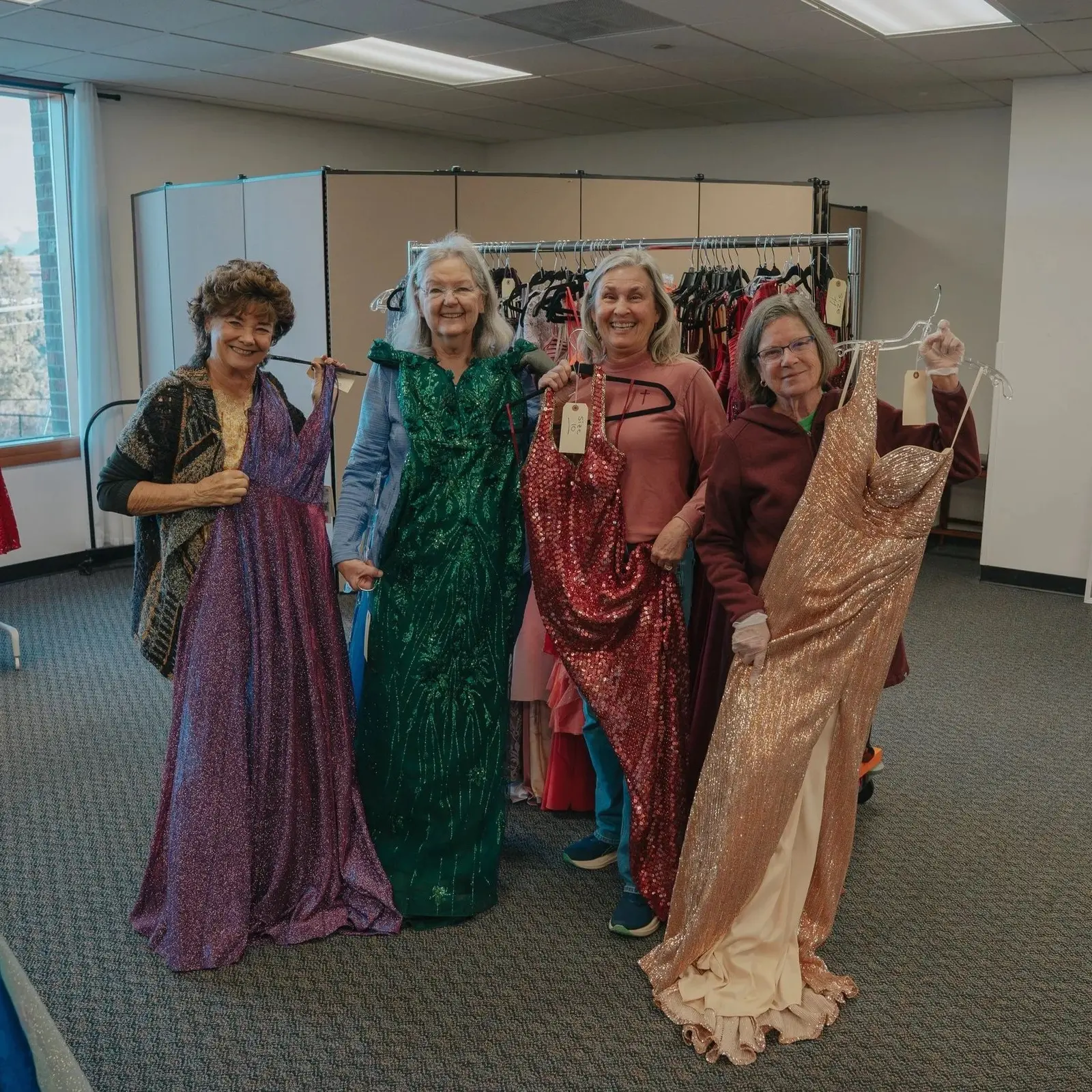 Four women hold up sparkling formal gowns in a room with a garment rack.