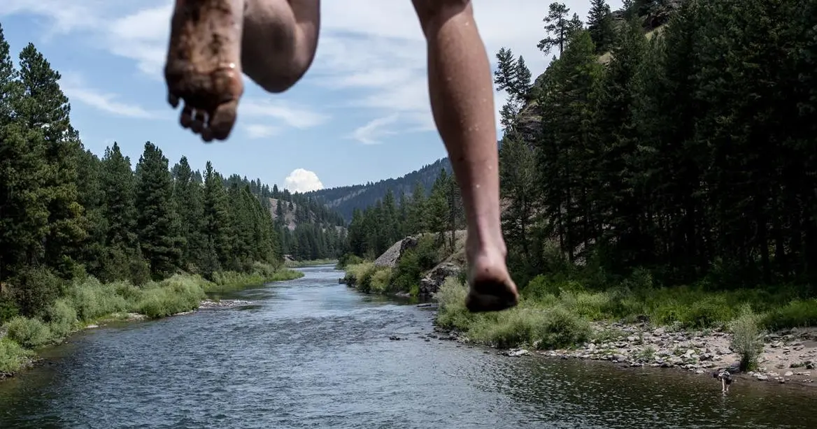A person jumps over a river surrounded by a forest.