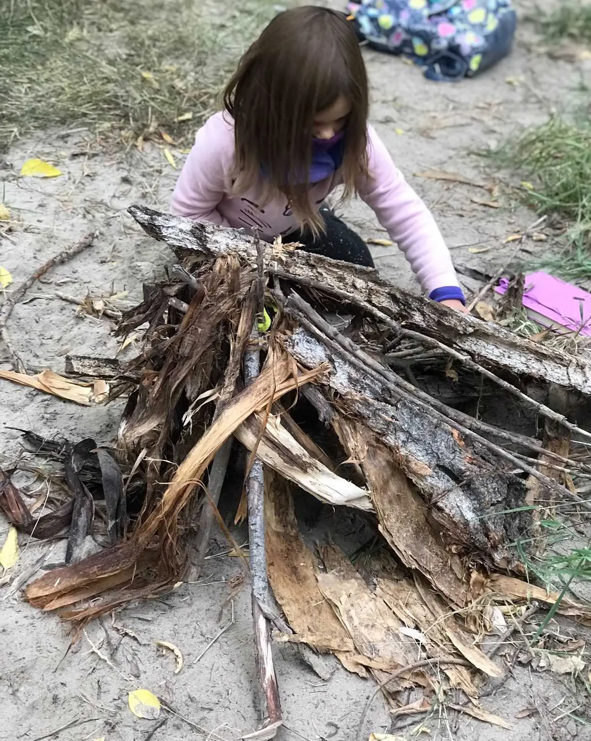 A child sits on the ground outdoors, arranging sticks and bark into a pile.