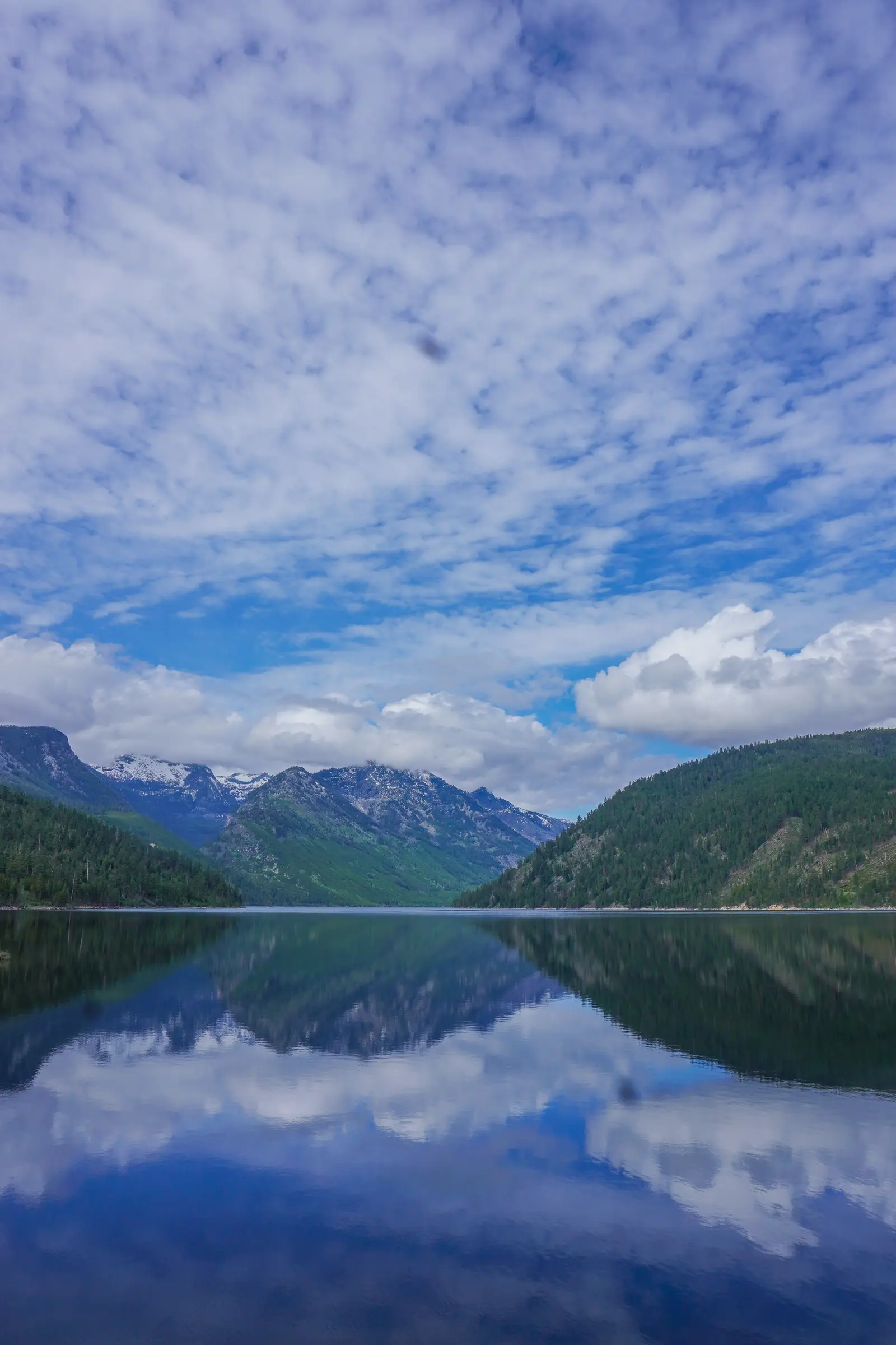 A calm lake reflects the surrounding forested mountains and cloudy sky.