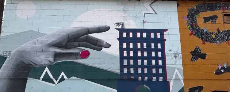 A mural on a cinder block wall depicts a giant hand reaching toward a building.