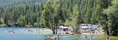 People enjoy swimming and relaxing at a crowded beach by a lake surrounded by a dense forest.
