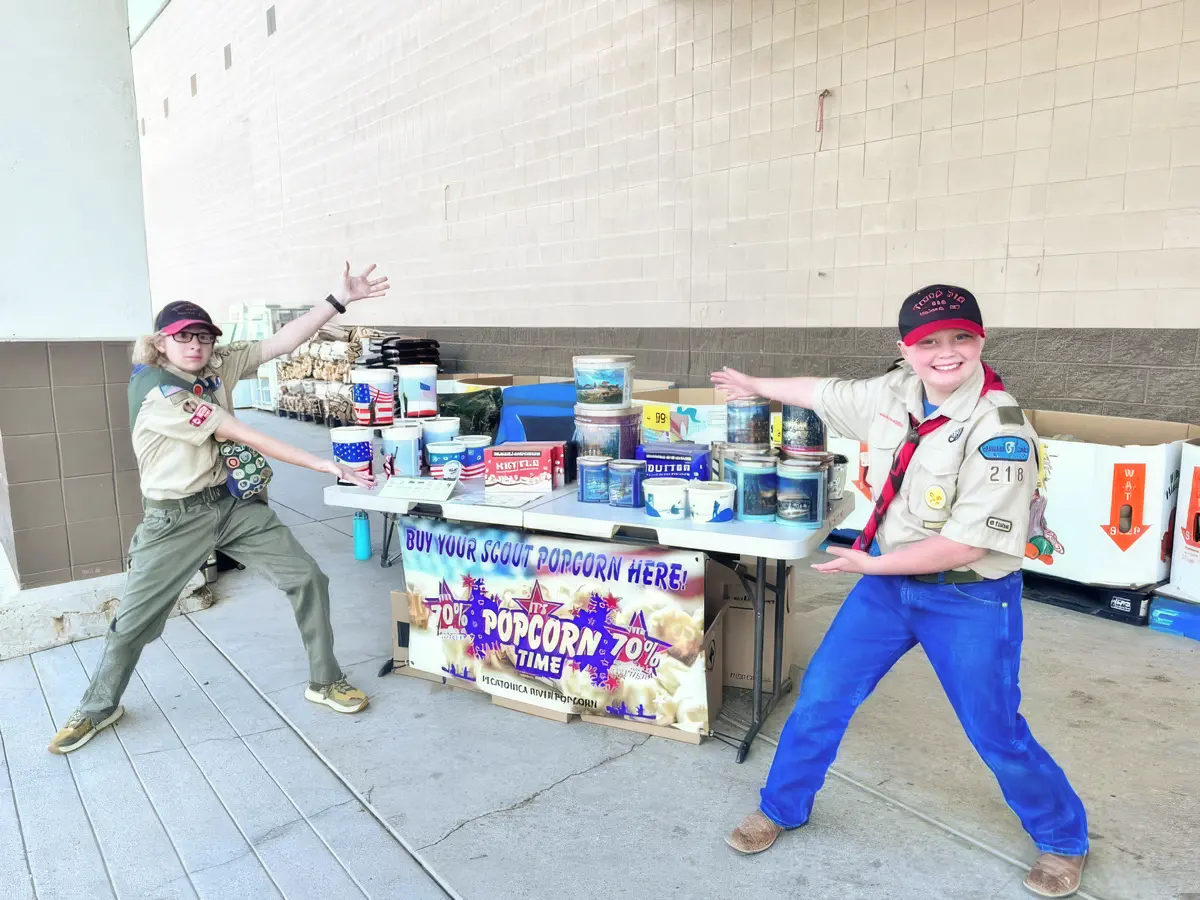Two children in scout uniforms pose in front of a popcorn sales table.