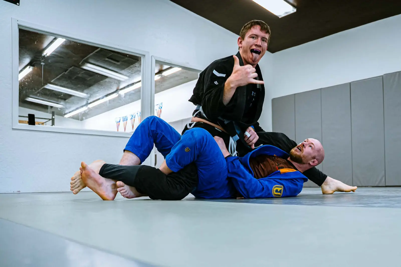 Two men in martial arts uniforms are grappling on a mat in a gym.