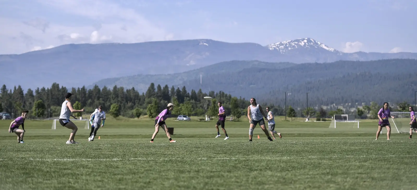 A group of young adults playing ultimate frisbee on a field with mountains in the background.