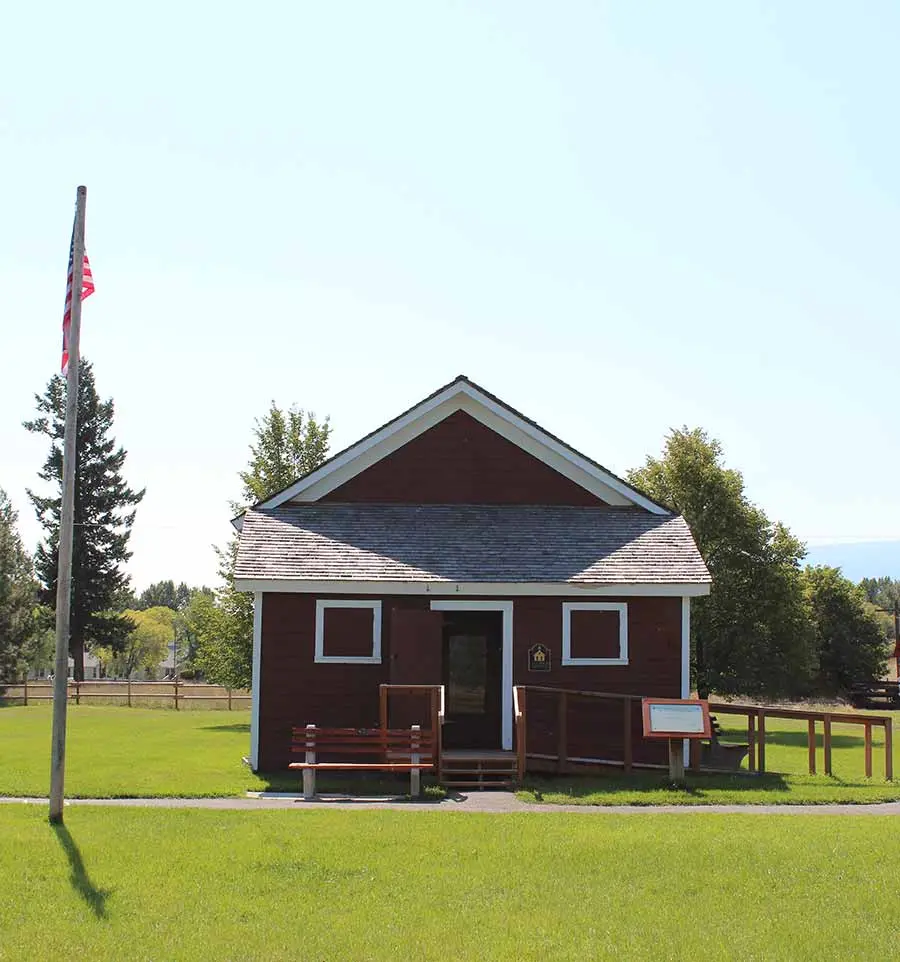 A small, dark red building with a shingled roof stands on a grassy lawn next to a flagpole.