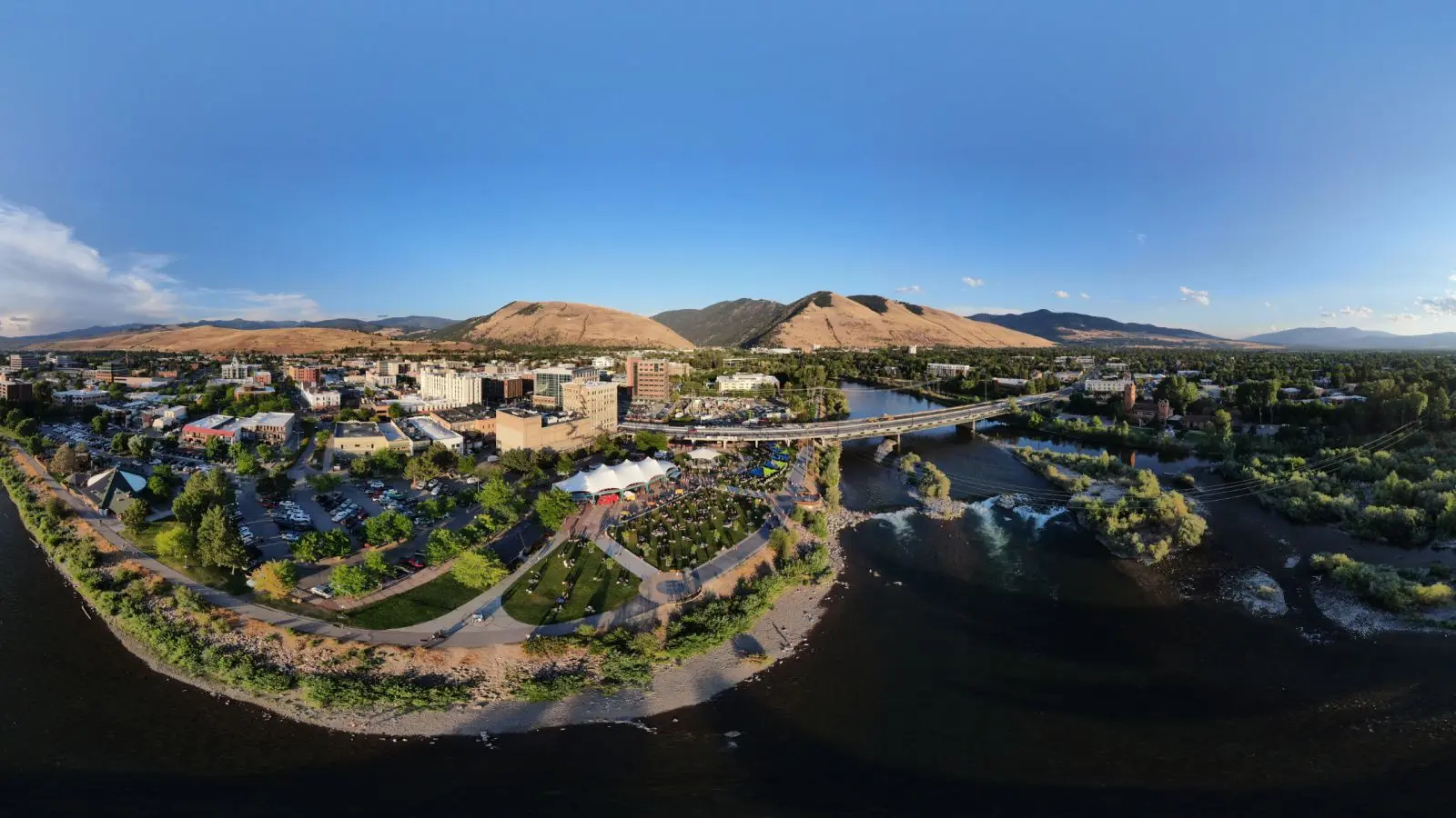 An aerial panoramic view shows a town situated along a river with mountains in the background.
