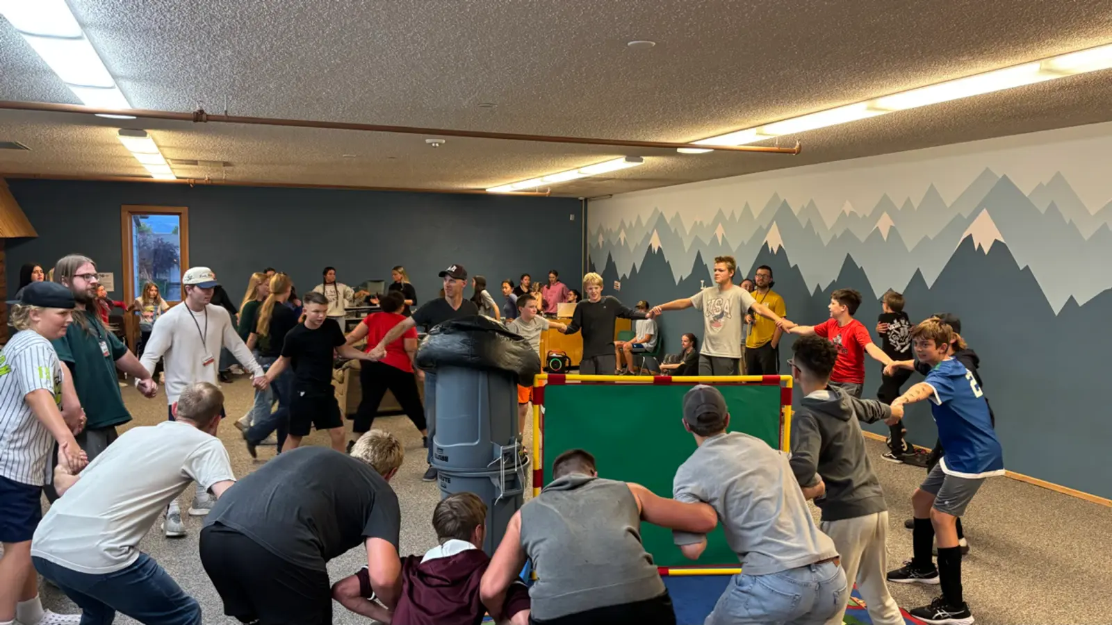 A large group of young people hold hands in a circle in a room with a mountain mural.