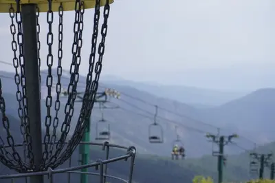 A disc golf basket stands in the foreground with a chairlift in the background over mountainous terrain.
