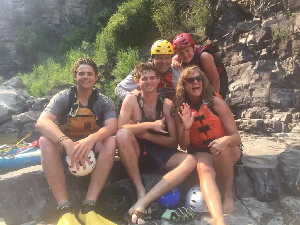 A family sits together on rocks while wearing safety gear for an outdoor water activity.