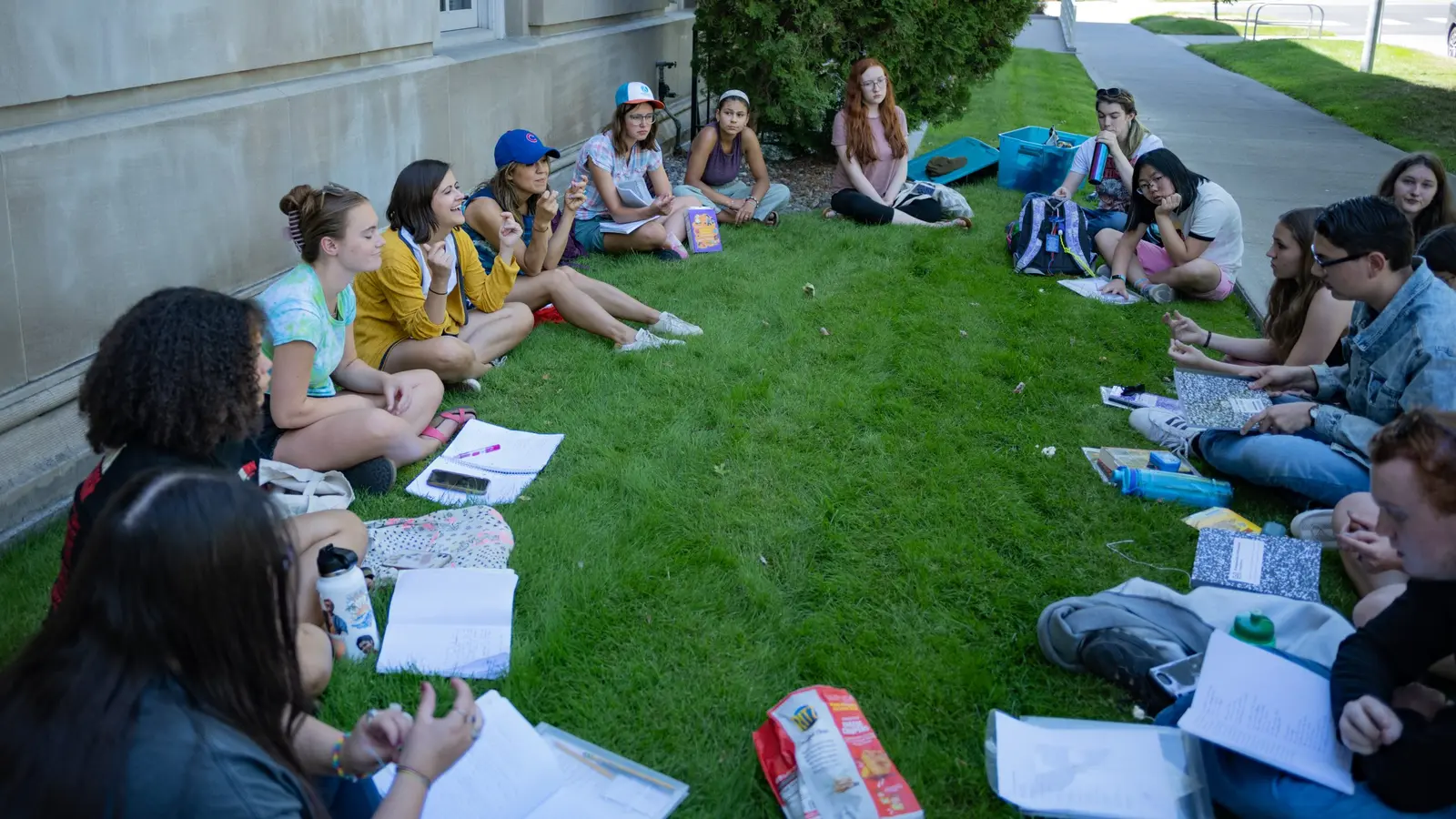 A group of young people sit in a circle on the grass outdoors with notebooks and papers.