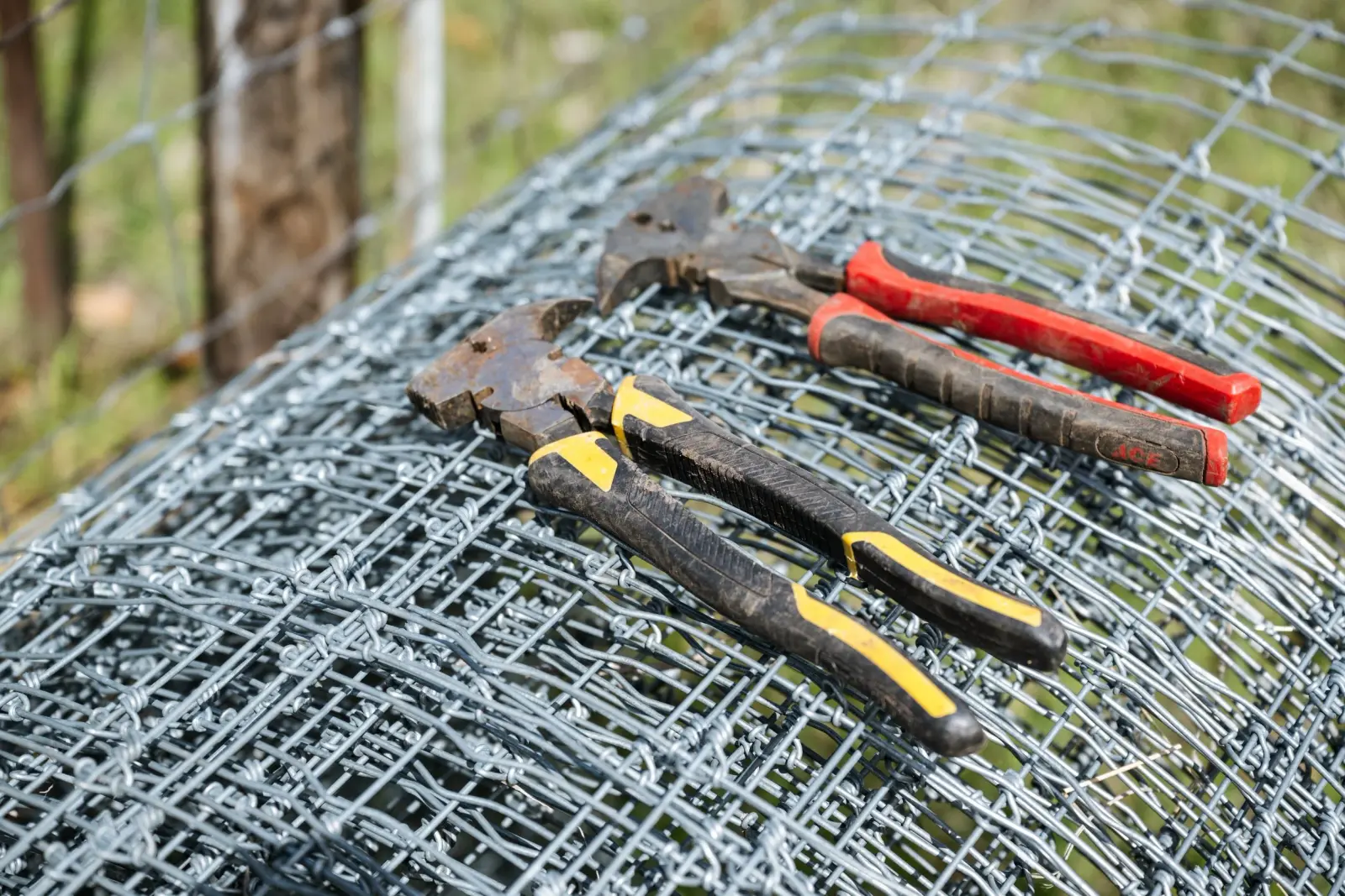 Two pairs of fencing pliers are resting on top of a roll of galvanized wire mesh fencing.