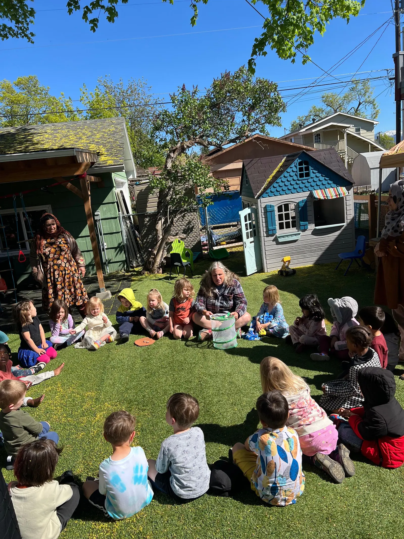 A group of children sitting in a circle on the grass outside, listening to a woman in the center who is showing them something in a bag. There are colorful houses, a tree, and a blue sky in the background.
