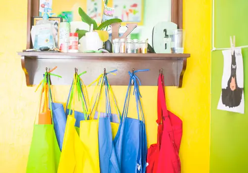 A shelf with supplies and colorful aprons hanging below it against a bright yellow and green wall.