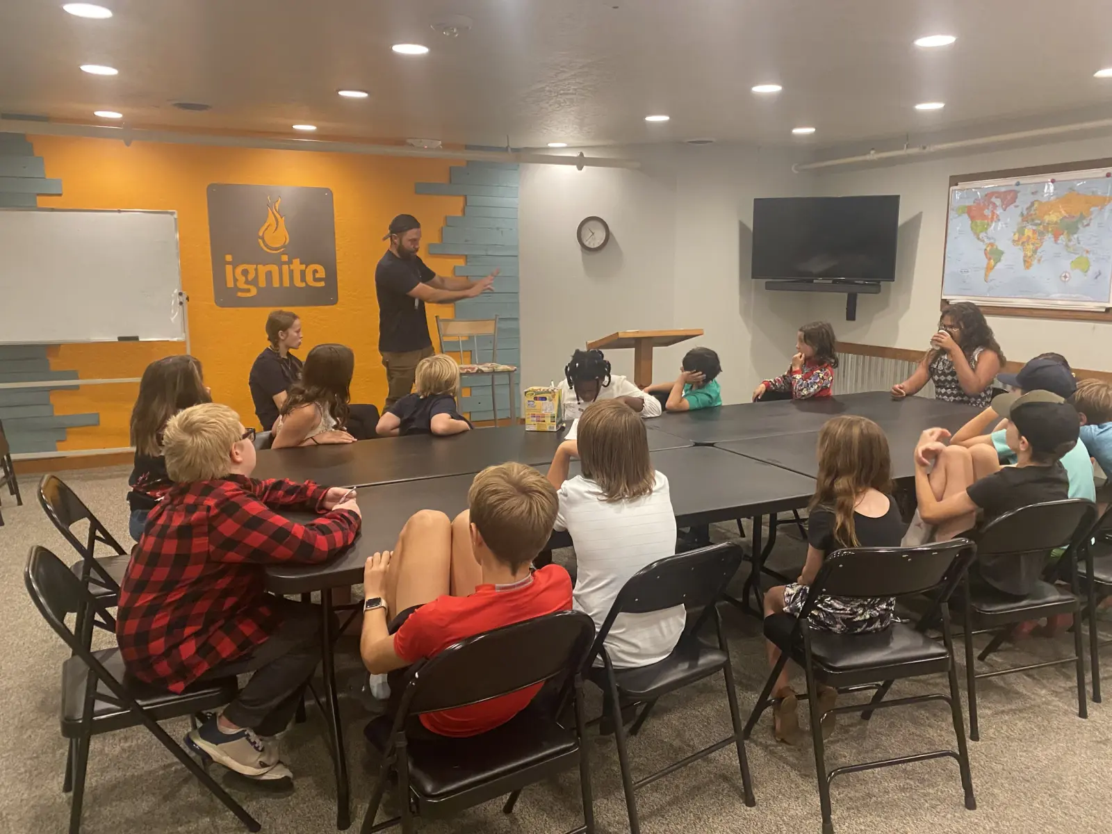 A man leads a group of children seated around tables in a room with an orange wall featuring an ignite logo.