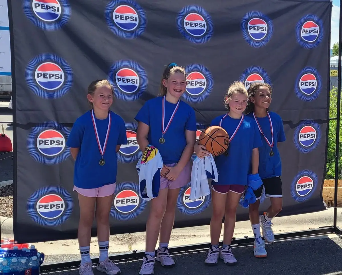 Young girls standing with medals and basketball in front of a Pepsi banner.