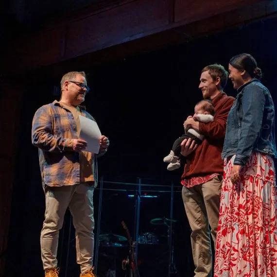 A man on stage holding a paper speaks to a couple holding a baby.