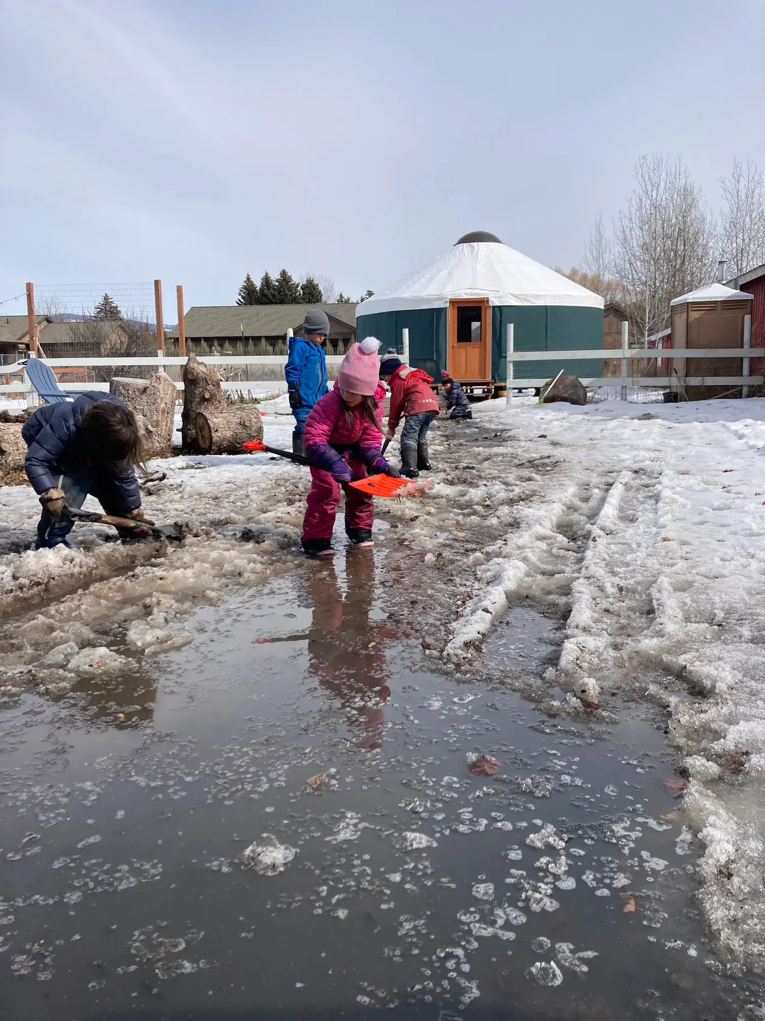 Children play with shovels in a melting snowy yard.