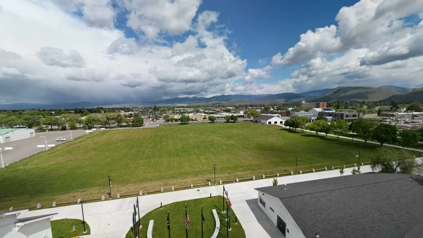 A high-angle view shows a large grassy field surrounded by buildings and parking areas under a cloudy sky.