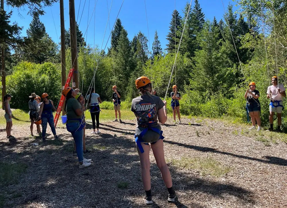 A group of young people wearing harnesses and helmets participate in an outdoor rope course activity.