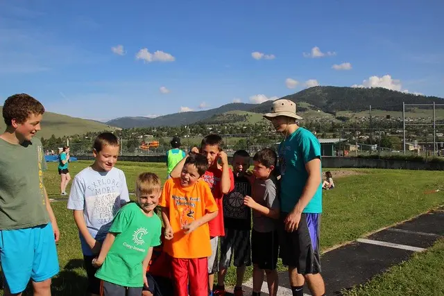 A group of children and an instructor stand together on a grassy field in front of a mountain landscape.