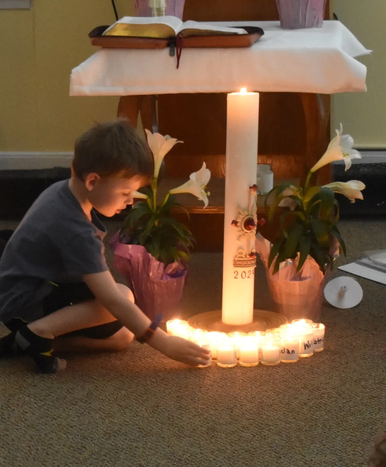 A young boy kneels on the carpet to touch one of many small votive candles arranged around a large pillar candle.
