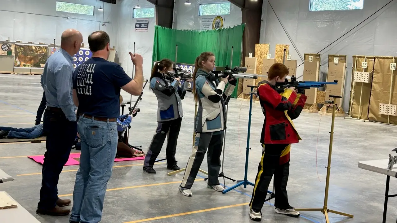 Young shooters practice with rifles at an indoor shooting range under the supervision of instructors.