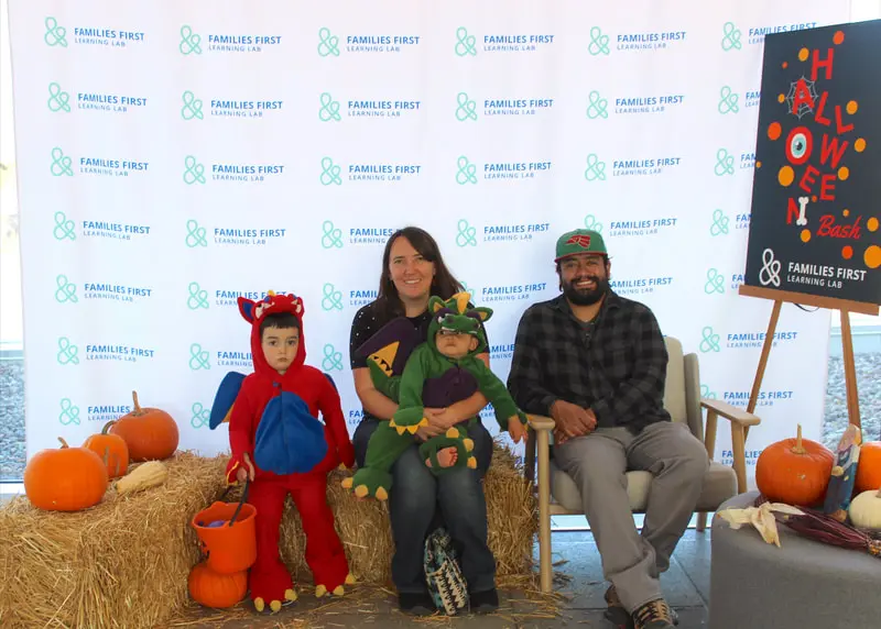 A family poses for a photo in Halloween costumes in front of a branded backdrop with pumpkins.