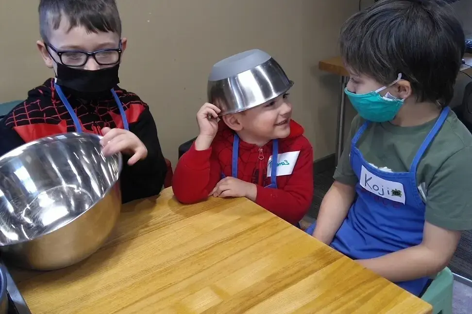 Three children wearing aprons and masks sit at a wooden table, with one child wearing a metal bowl on their head.