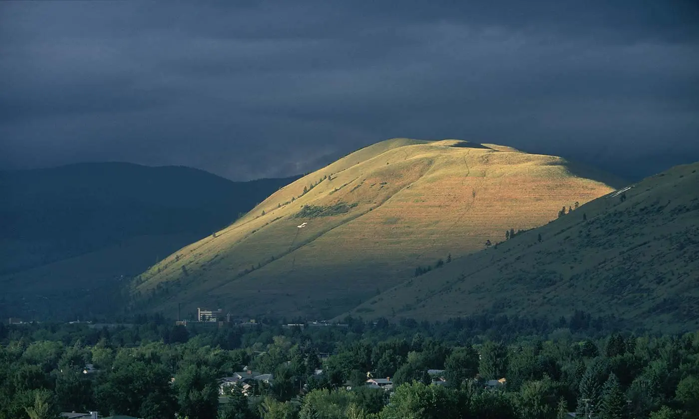A brightly lit grassy hill contrasts with dark, stormy clouds above a town.