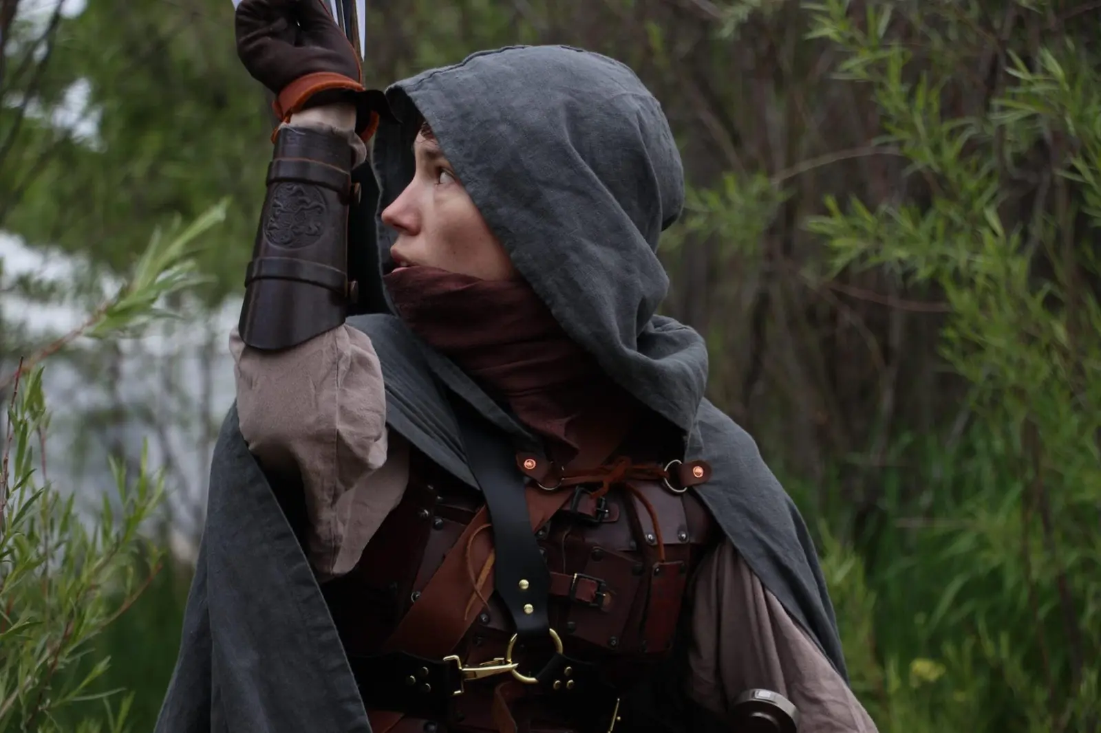 A person dressed in fantasy attire with a hooded cloak reaches up to hold an arrow.