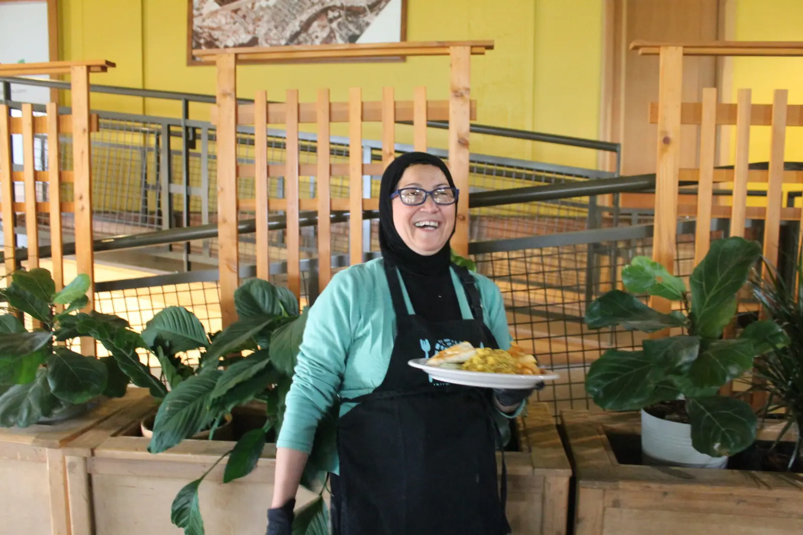 A smiling woman wearing an apron and hijab holds a plate of food.