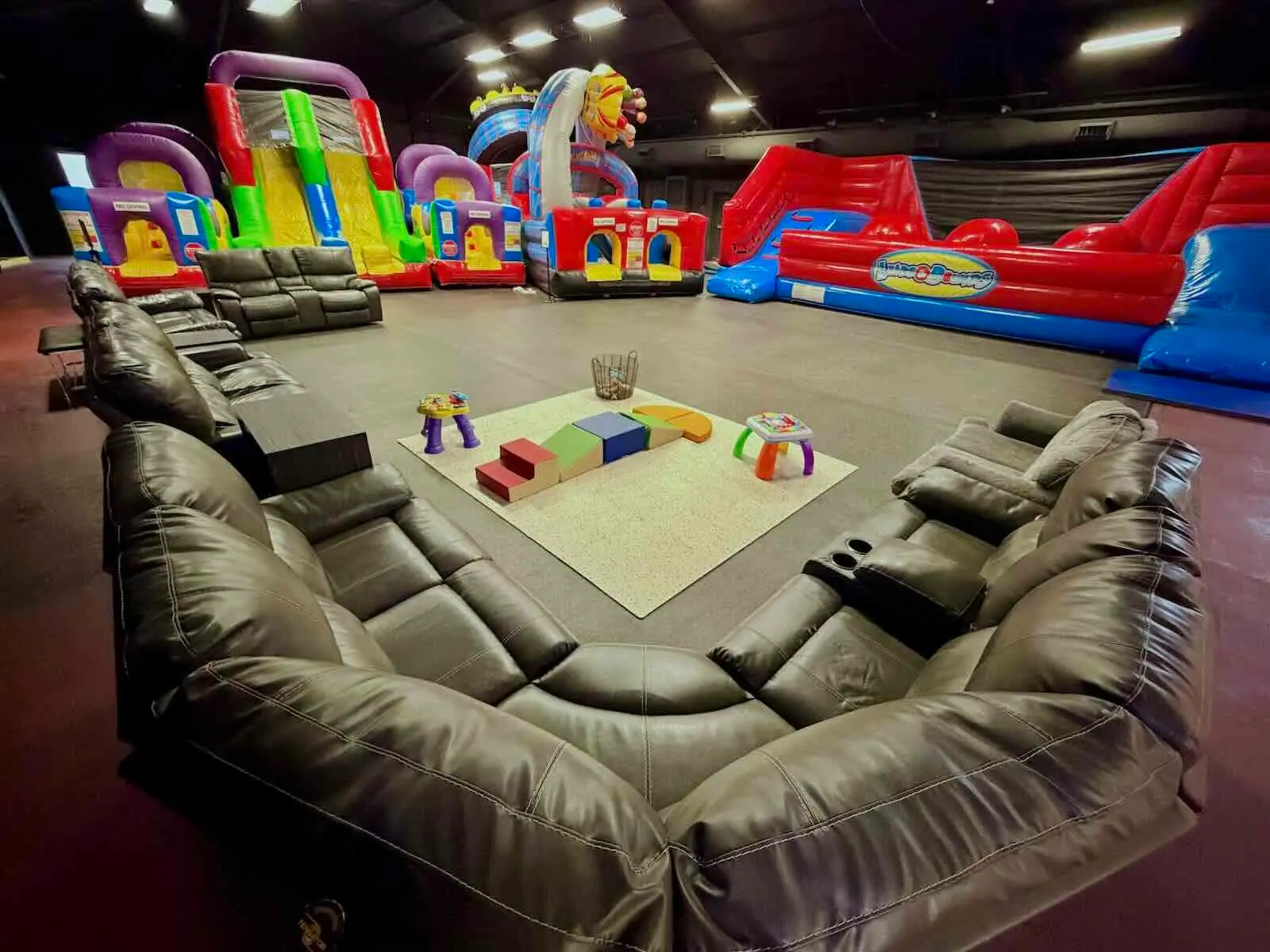 Indoor play area with colorful inflatable slides and bounce house, surrounded by brown leather couches, a central play mat with toys, and a dark ceiling.