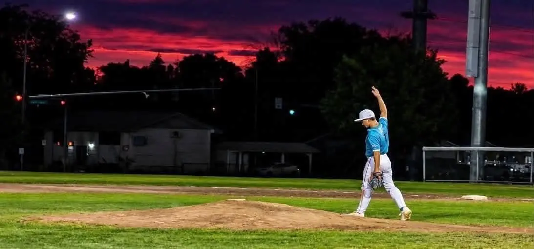 A baseball player walks off the mound against a dramatic sunset background.