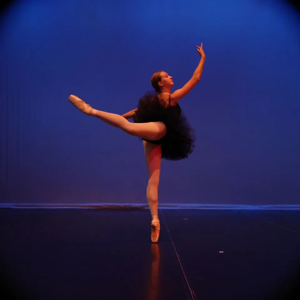 A ballerina in a black tutu performs a dance move on a stage.