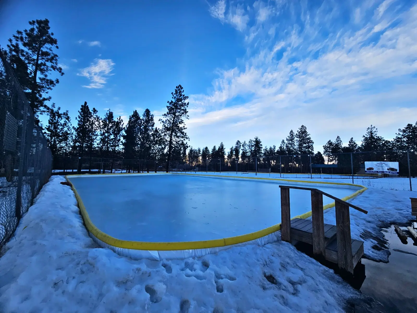 An outdoor ice skating rink is surrounded by snow and trees under a blue sky.
