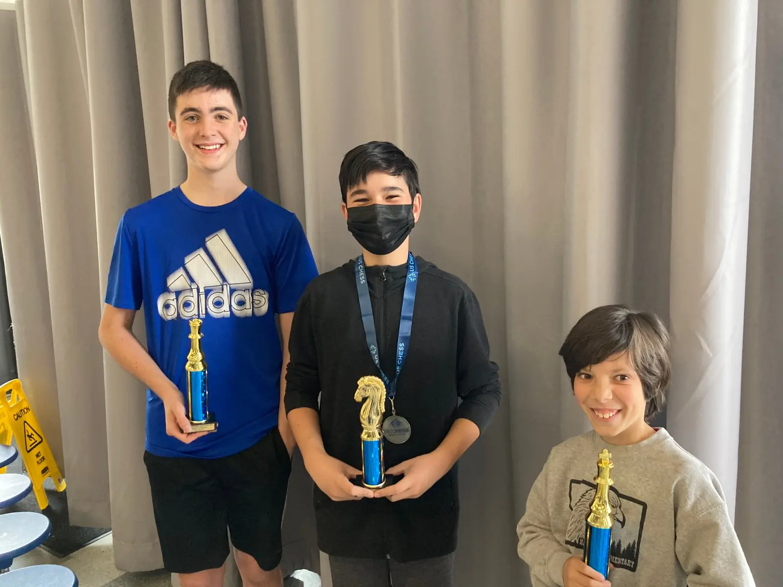 Three young people hold trophies and stand in front of a curtain.