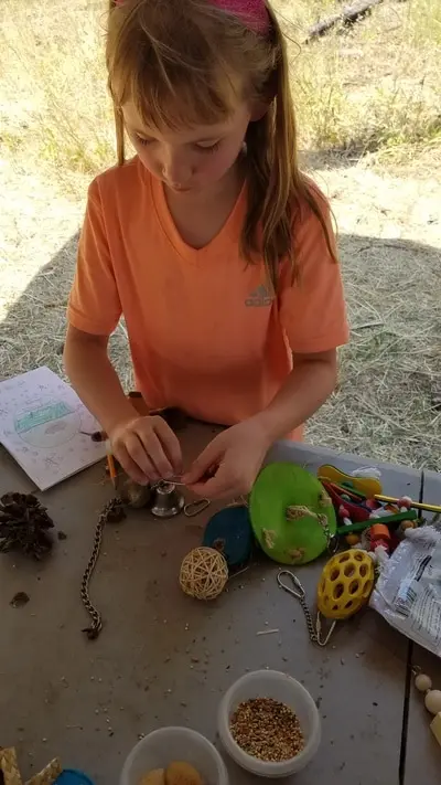 A young girl works on a craft project with various materials at a table outdoors.